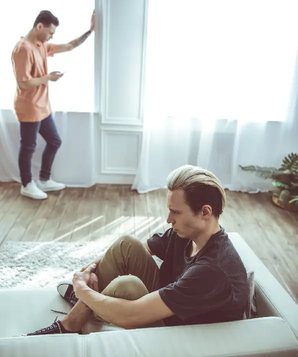 Toned portrait of unhappy guy with dyed hair sitting on couch while his boyfriend standing near window and using smartphone Toned portrait of unhappy guy with dyed hair sitting on couch while his boyfriend standing near window and using smartphone