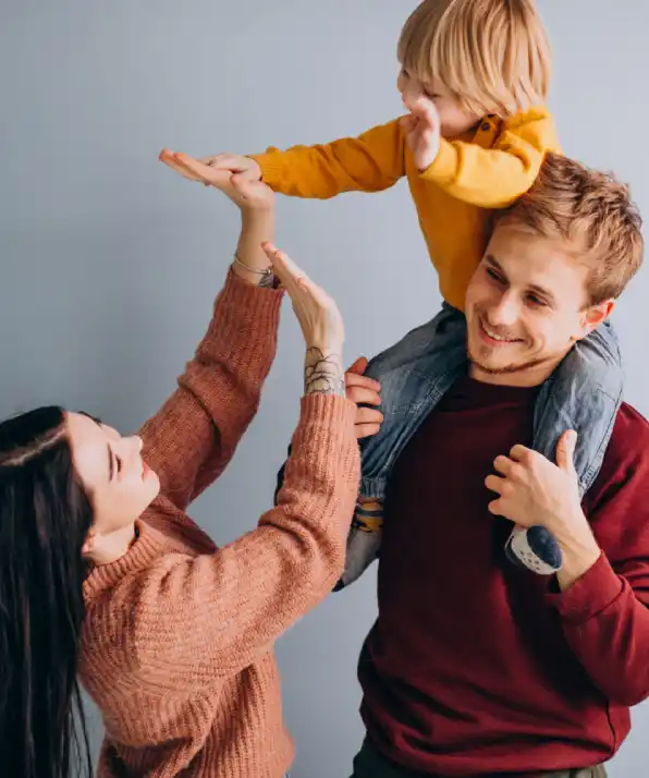A young child in a yellow shirt is playfully lifted by a person sitting on another's shoulders, against a soft gray background
